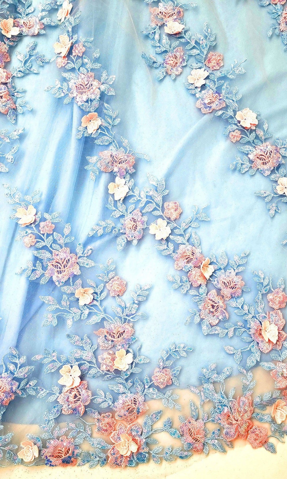The Quinceanera Dress CHF3117 by Chicas features a light blue sheer fabric beautifully adorned with pink and blue embroidered floral patterns arranged diagonally, complemented by delicate 3D floral appliques that add dimension and eleganceโmaking it the perfect dreamy choice for a quinceaรฑera or tulle ballgown.
